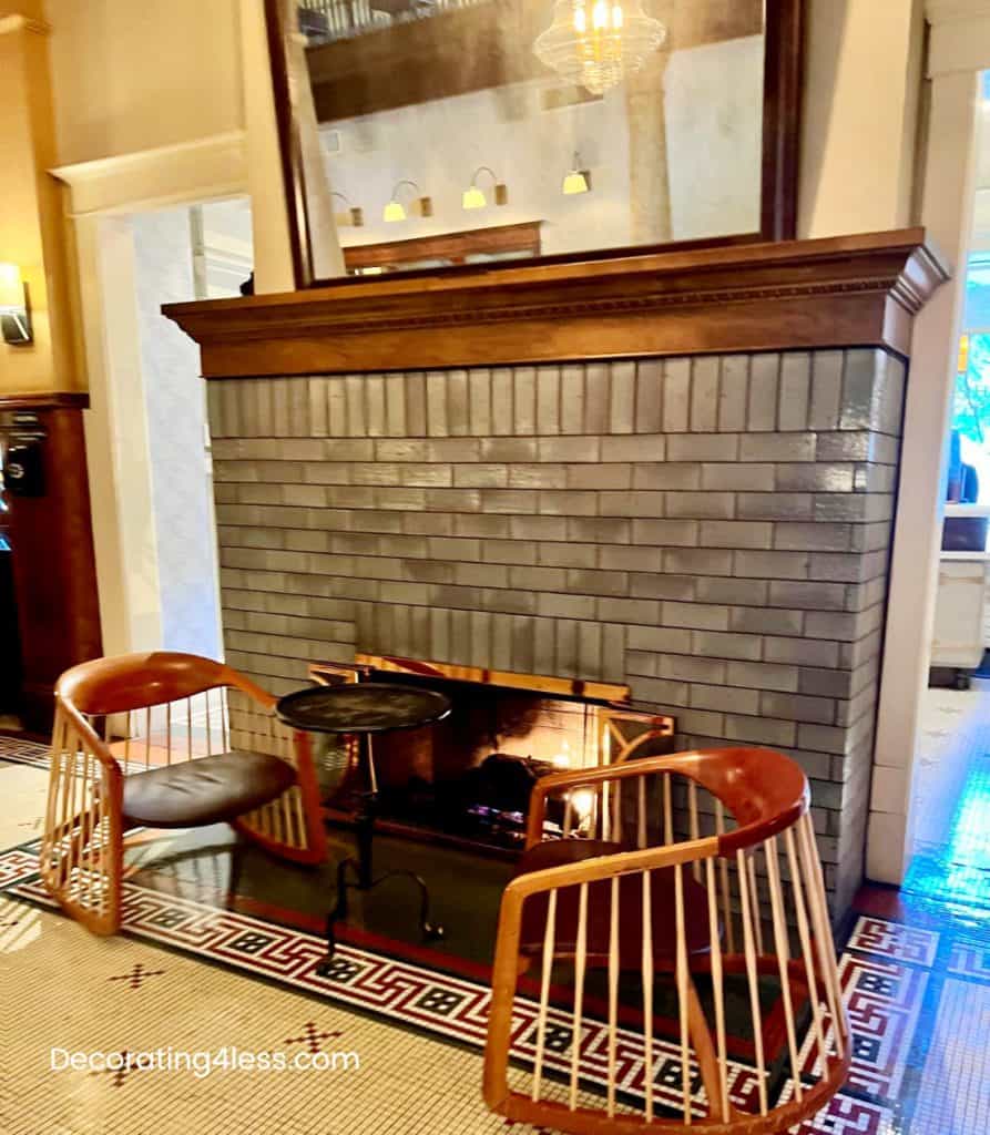 Dark gray brick fireplace with mirror, wooden mantel centerpiece in cozy living room decorated for fall, with two rattan chairs, a small black side table, and a decorative area rug. Perfect for home decorating on a budget.